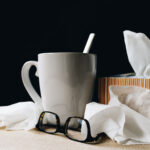 Taza de bebida caliente junto a pañuelos y gafas, símbolo de resfriado o cuidado en casa