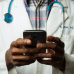 Médico revisando su teléfono móvil con un estetoscopio azul colgado del cuello.