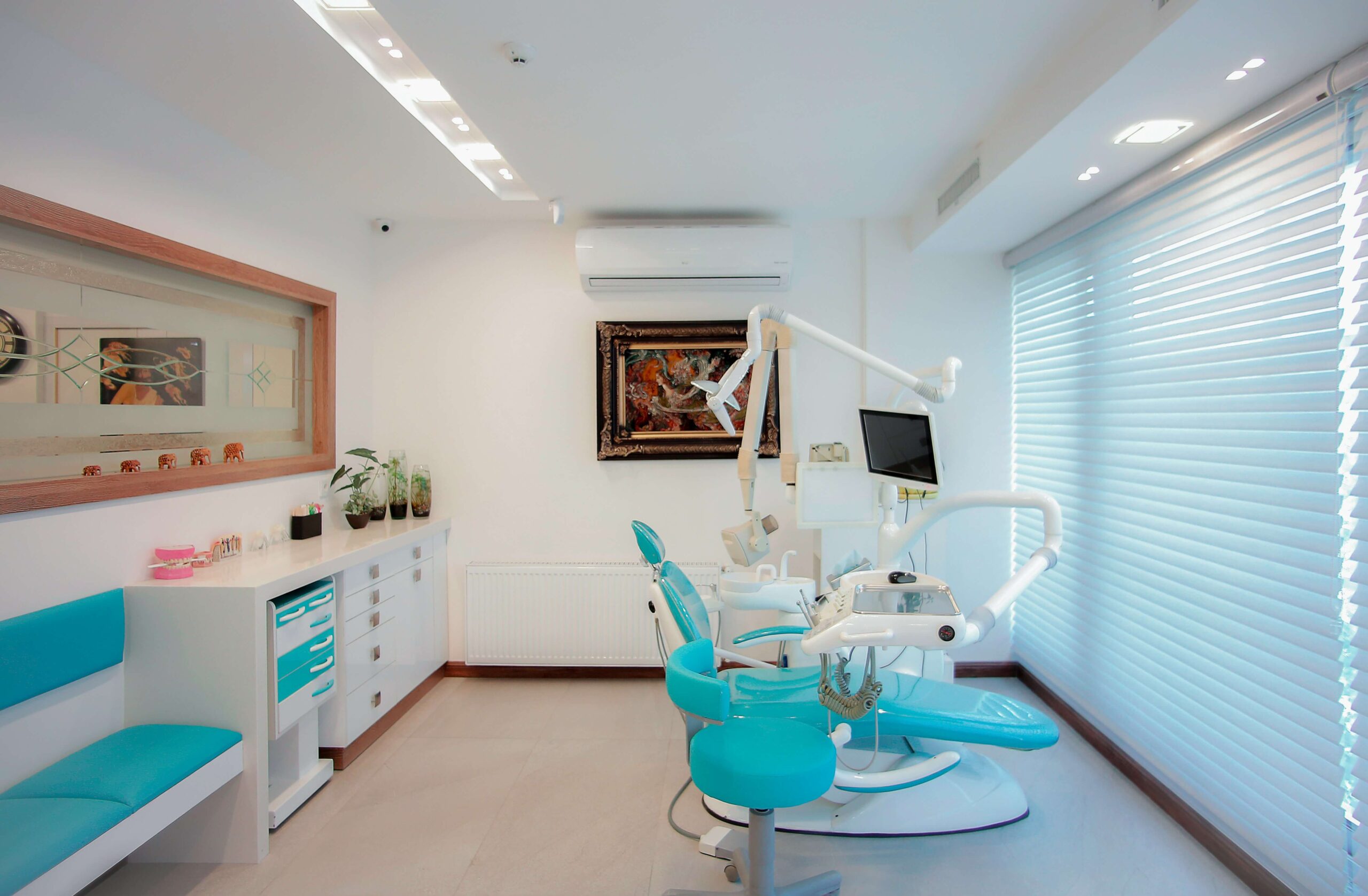 Interior de consulta dental con muebles en tonos turquesa, camilla de examen, equipo dental moderno y ventana con persianas blanca